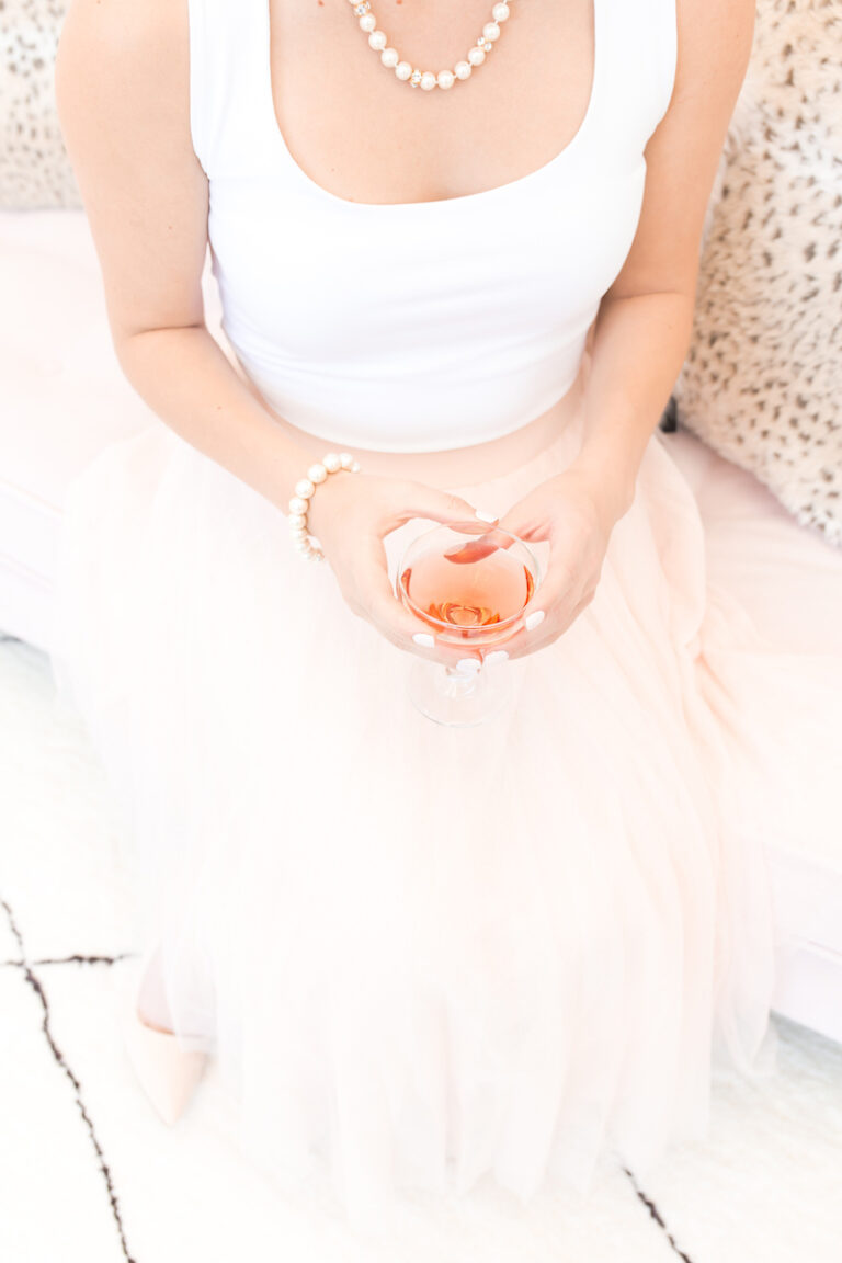 Woman dressed in classic pink skirt and white top wearing a pearl bracelet and pearl necklace and holding a pink beverage.