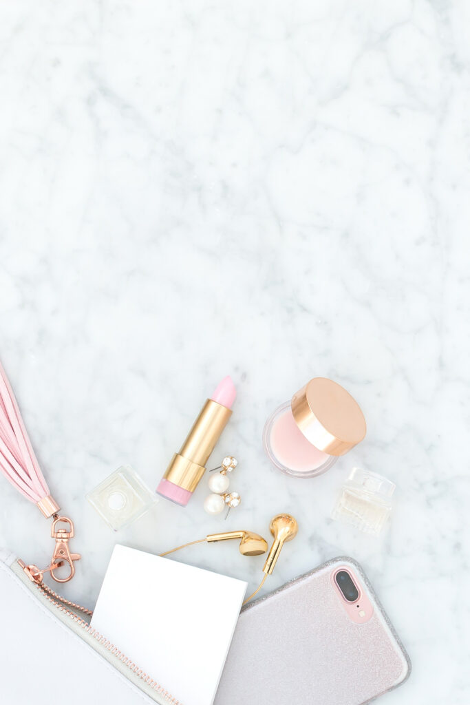 Pearl earrings, pink lipstick, pink makeup, purse with pink tassel, gold ear pods and pink-tinted iPhone arranged on classy marble background.